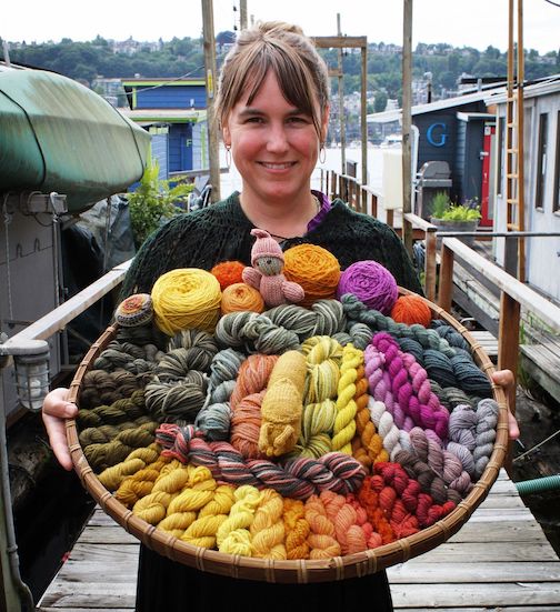 lissa Allen, stands holding forth a basket that is full of colourful wool dyed with mushrooms and lichens. There are yellows, greens, pinks, oranges, and everything in between!  Photograph courtesy of Megan Hodde. To find out more, click here.